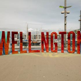 The Well_ngton Sign is located at the Wellington Harbourfront Walk, with a metallic red facade reflecting light onto the cement in the foreground.