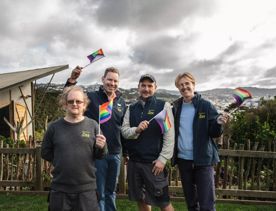 Four Wellington Zoo staff members stand together smiling and waving LGBTQIA+ flags in front of a wooden fence with greenery, hills and an overcast sky in the background.