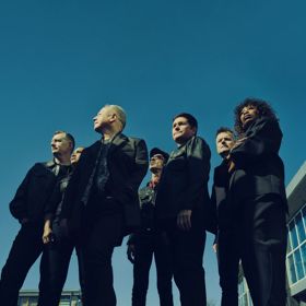Simple Minds band members standing outdoors in dark suits against a clear blue sky and modern building backdrop.