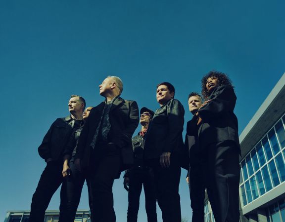 Simple Minds band members standing outdoors in dark suits against a clear blue sky and modern building backdrop.