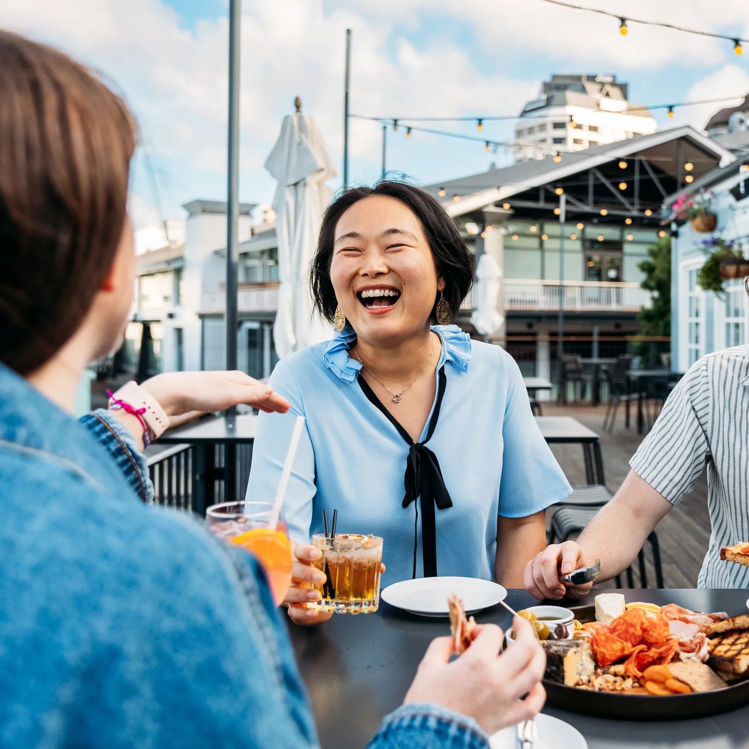 Three people sit at a bar table at Foxglove Bar on the Wellington Waterfront. They are all drinking a cocktail and a charcuterie board sits between them.
