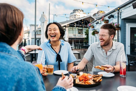Three people sit at a bar table at Foxglove Bar on the Wellington Waterfront. They are all drinking a cocktail and a charcuterie board sits between them.