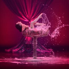 Burlesque performer Dita Von Teese splashes soapy water in an oversized, ornate martini glass on a pink stage.