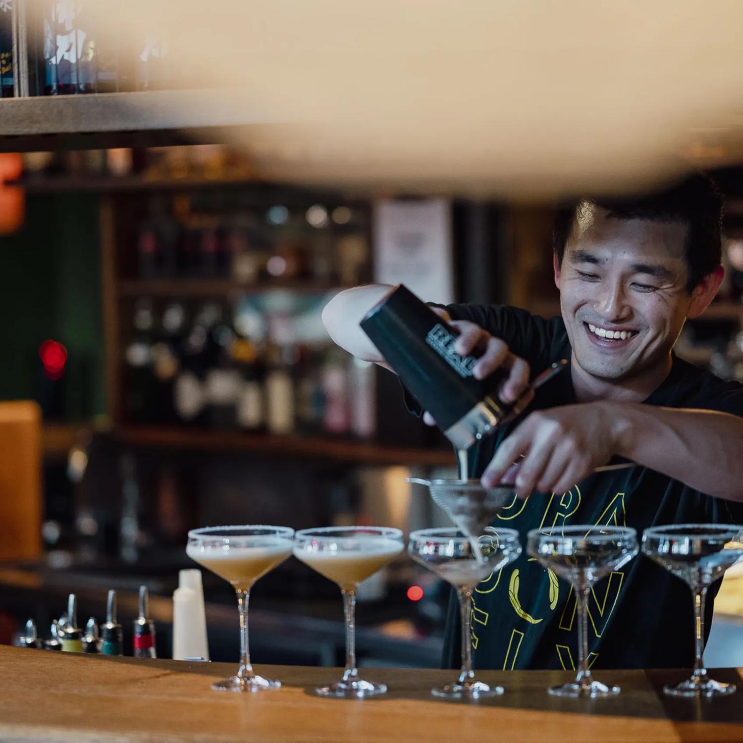 A member of the Dragonfly bar staff stands behind the bar pouring a cocktail mixer into five glasses. Behind are shelves full of bottles.