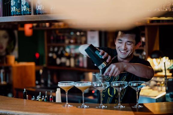 A member of the Dragonfly bar staff stands behind the bar pouring a cocktail mixer into five glasses. Behind are shelves full of bottles.