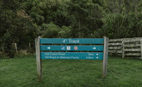 The sign at the bottom of the 4 degrees Track in Belmont Regional Park. The sign reads '4 Degrees Track, Old Coach Road 3km and Hill Road 4.5km.' There are symbols that show walkers and bikes are welcome, but no horses.