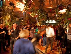 A party with heaps of people at the St James Theatre. The space is decorated with handing plants and amber lighting. 