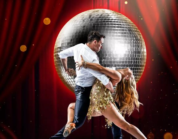 Two dancers are doing a romantic pose on a stage farmed with red curtains and a massive disco ball behind them.