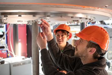 Two OpenStar employees wearing orange hardhats maintain a piece of metal machinery.