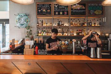 Two bartenders, one shaking a cocktail and the other pouring a pint, wear black 'Burger Liquor' teeshirts while working.