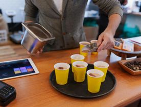 A staff member at Wellington Chocolate Factory sprinkles chocolate dustings onto five yellow caps on a tray, which have hot chocolate in them.