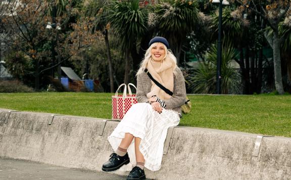 Emma Smith, a Wellington-based artist, is seated in a park wearing a blue knit hat, a beige scarf, brown-grey jumper and white skirt. 