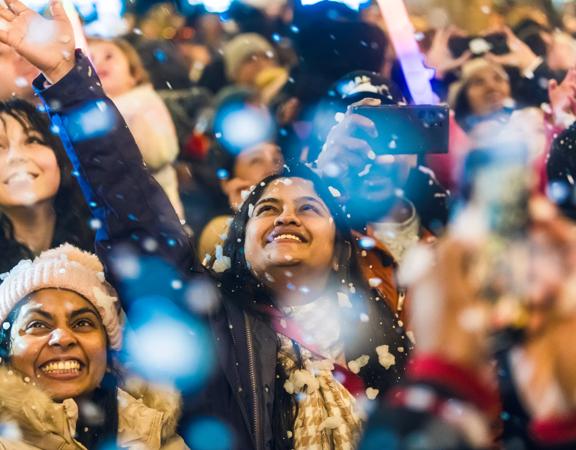 A crowd of smiling people in winter wear enjoying the falling snow at Greytown Festival of Christmas.