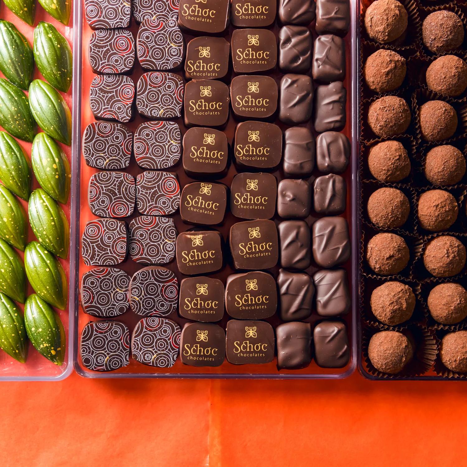 Three trays clear plastic of artisanal chocolates from Schoc Chocolates shop on a red-orange surface. 