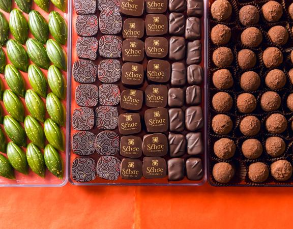 Three trays clear plastic of artisanal chocolates from Schoc Chocolates shop on a red-orange surface.