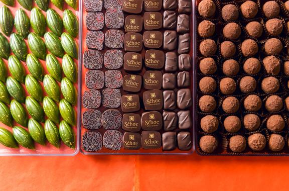 Three trays clear plastic of artisanal chocolates from Schoc Chocolates shop on a red-orange surface.