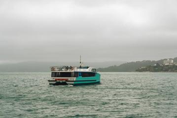 The teal-coloured Ika Rere electric ferry in the Wellington harbour on a cloudy, foggy day.