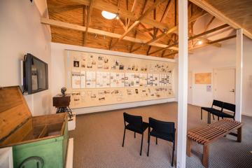 Inside Nairn Street Cottage in Wellington. The room has white walls, a pitched wooden roof, a bench, four black chairs, and a wall displaying images and historical information.