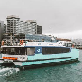 The teal-coloured Ika Rere electric ferry leaves its berth on the Wellington waterfront to head out into the harbour. The Wellington skyline is behind it.
