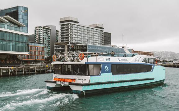 The teal-coloured Ika Rere electric ferry leaves its berth on the Wellington waterfront to head out into the harbour. The Wellington skyline is behind it.