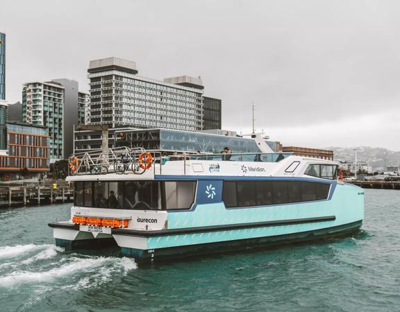 The teal-coloured Ika Rere electric ferry leaves its berth on the Wellington waterfront to head out into the harbour. The Wellington skyline is behind it.