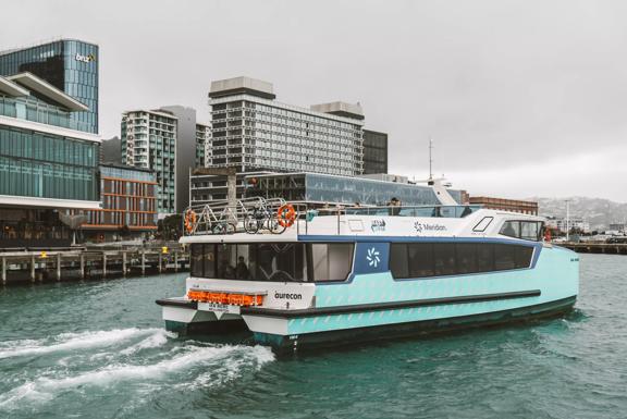 The teal-coloured Ika Rere electric ferry leaves its berth on the Wellington waterfront to head out into the harbour. The Wellington skyline is behind it.