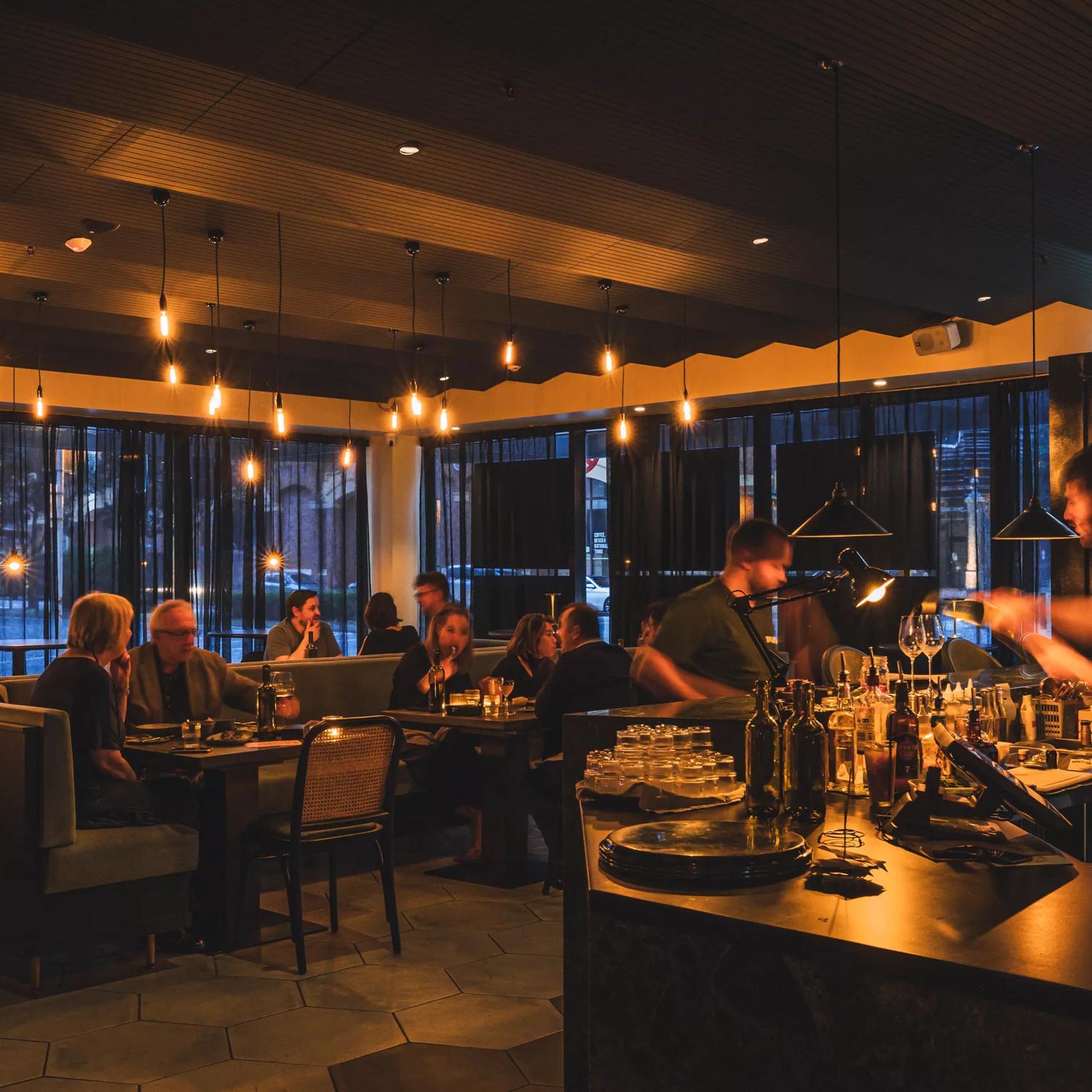 The interior of Plonk under low light, customers sit in the background enjoying their meals and bar staff serve drinks in the front.