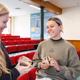 Two smiling people, each holding a be happy chocolate product, are inside a lecture hall at Victoria University. 