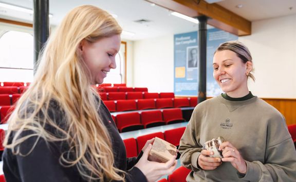 Two smiling people, each holding a be happy chocolate product, are inside a lecture hall at Victoria University.