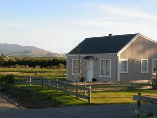 Small accommodation house with tree in front and fence surrounding, and hills in background.