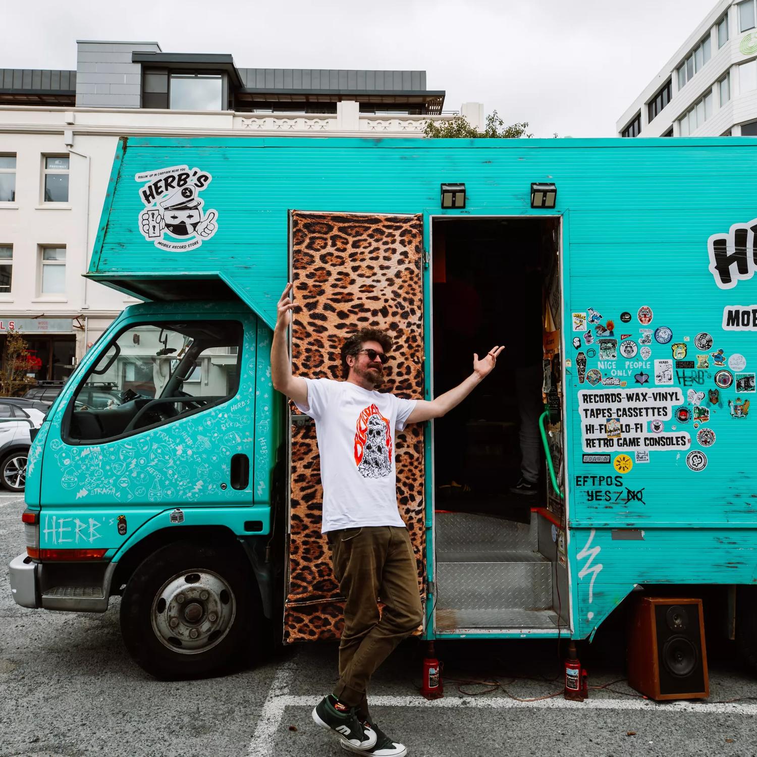 Ben James, owner of Herb's Mobile Record Store, stands outside his 2004 Mitsubishi Canter truck. The moving record shop is painted blue-green and decorated with stickers.