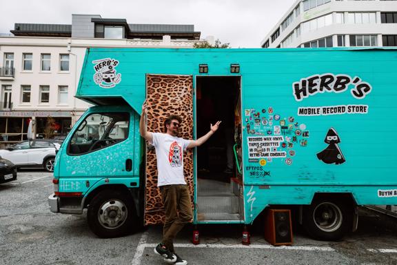 Ben James, owner of Herb's Mobile Record Store, stands outside his 2004 Mitsubishi Canter truck. The moving record shop is painted blue-green and decorated with stickers.