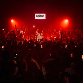 Crowd gathered at a live music event under red stage lighting, with audience members raising hands and holding up phones.