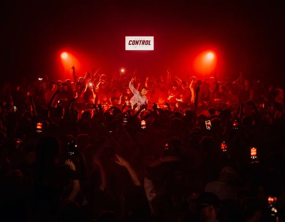 Crowd gathered at a live music event under red stage lighting, with audience members raising hands and holding up phones.