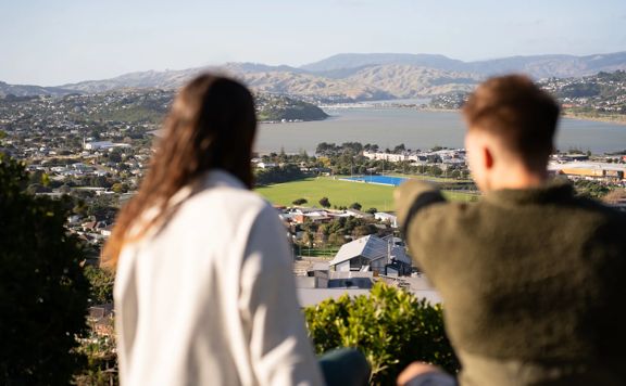 Two people sit on a bench at Rangitoto Lookout. One points down at the town below.