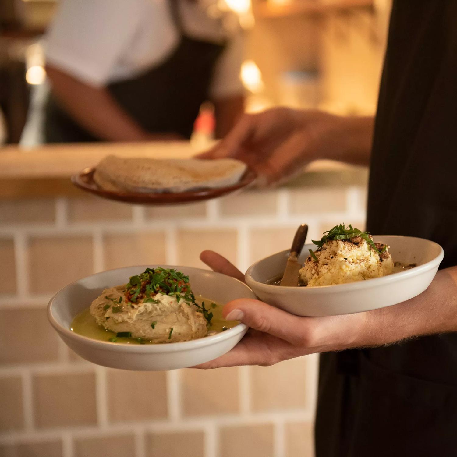 A waiter wearing a white T-shirt and dark green apron is holding two plates of hummus in one hand and a plate of pita in the other at Kisa, a Mediterranean restaurant in Te Aro, Wellington.