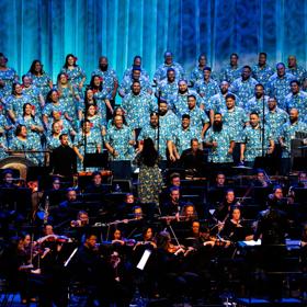 A large choir wearing matching blue shirts sings against a blue backdrop. In front of them is a large orchestra, mostly dressed in black, and playing instruments.