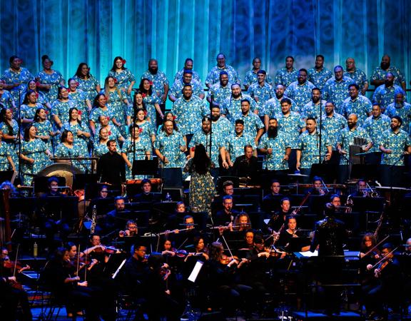 A large choir wearing matching blue shirts sings against a blue backdrop. In front of them is a large orchestra, mostly dressed in black, and playing instruments.