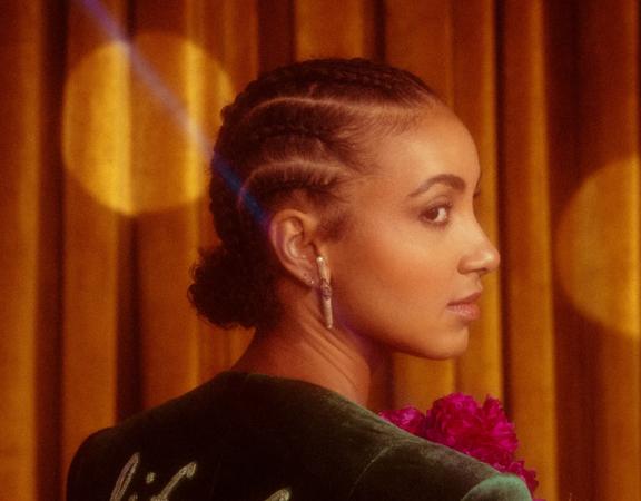 Esperanza Spalding stands facing a velvet curtain, holding a red bouquet and looks over her right shoulder.