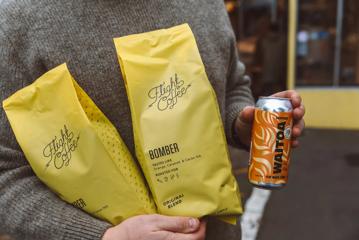 A person carries two yellow bags of Bomber Flight Coffee in one arm and a tall can of Waitoa 'Earlybird' stout.