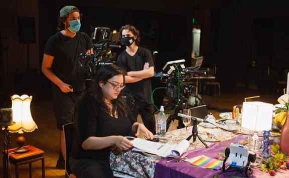 Pachali Brewster at a table looking at a script, while a camera crew stands behind.