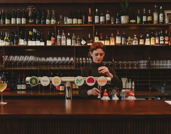 The Bar staff behind the bar at The Ram pouring craft beer. Theres a large shelf of wine and spirits bottles behind them.