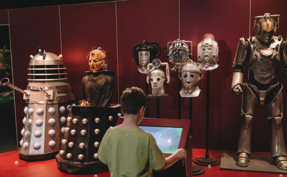 A child looks at a touch screen in front of a row of monsters from Doctor Who. The monsters include a Dalek, a Cyberman, and 5 detached Cybermen heads.