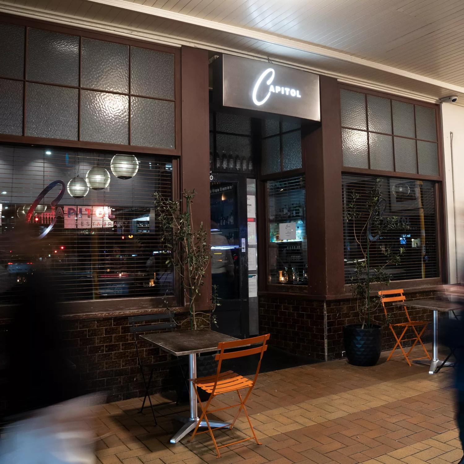 The front facade of Capitol Restaurant in Te Aro Wellington. The door is centered between two windows with dark brown trim and an illuminated sign suspended above. 