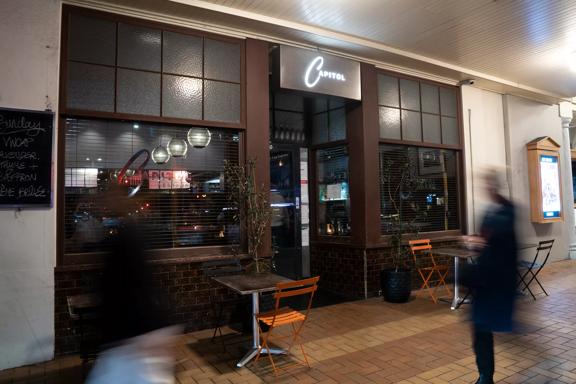 The front facade of Capitol Restaurant in Te Aro Wellington. The door is centered between two windows with dark brown trim and an illuminated sign suspended above. 