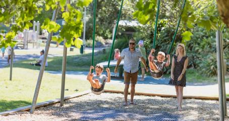 Two adults push two kids on swings at a playground on a sunny day.