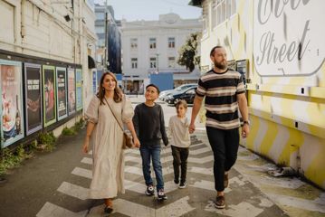 A family of four walks together down a city street, with buildings, posters, and a painted wall visible along the walkway.