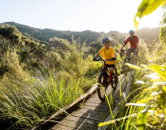A child and an adult bike along a wooden boardwalk amongst bush in the Wainuiomata bike park.