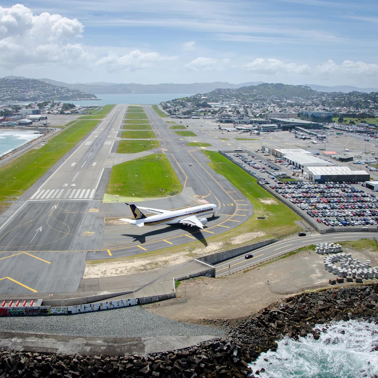 A drone shot of the runway at Wellington International Airport with a plane on the runway, a parking lot filled with cars located at Stewart Duff Drive, Rongotai with Hataitai, Evans Bay and Miramar seen in the background. 