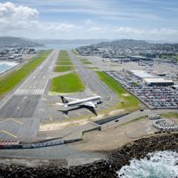 A drone shot of the runway at Wellington International Airport with a plane on the runway, a parking lot filled with cars located at Stewart Duff Drive, Rongotai with Hataitai, Evans Bay and Miramar seen in the background.